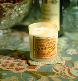 Lit Gardenia & Oud candle with ornate label rests on a woven coaster atop a floral tablecloth with crystal and a glass bottle in the background