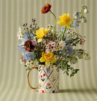 White porcelain mug with colorful floral designs and gold details holds a bouquet of flowers against a green and white striped backdrop