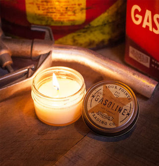 Lit Gasoline jar candle with lid removed is staged on a wooden surface with a gas pump nozzle and red gas cans in the background