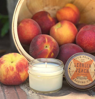 Small Georgia Peach scented jar candle with tin lid removed is staged with a basket of peaches on a rustic surface