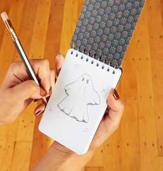 Models hands hold a pencil and a small spiral-bound notepad with decorative interior cover and ghost sketch on the first white page