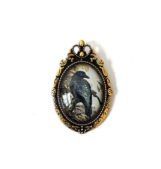 Oval brooch features illustration of a crow under clear glass in an ornate antiqued gold fame