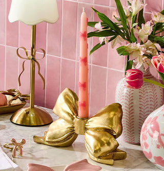 Pink taper candle accented with dark pink bows is held in a sculptural gold bow on a marble tabletop with other pink and bow-shaped items against a pink tiled backdrop