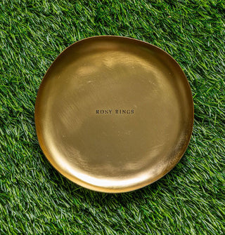 Slightly irregular round gold dish resting on astroturf is imprinted with the words, "Rosy Rings"