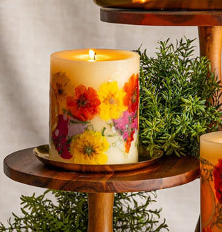 A lit single-wick pillar candle with colorful flowers embedded in its wax is situated in a gold platter on a wooden pedestal flanked by shrubs and other candle pedestals