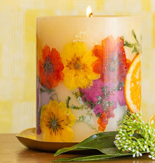 A lit single-wick pillar candle with colorful flowers and leaves embedded in its wax is situated on a gold platter and staged with a flower and leaves in the foreground
