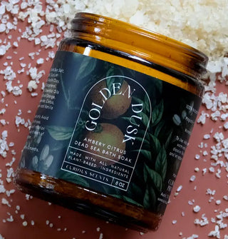 Amber jar of Golden Dusk Ambery Citrus Dead Sea Bath Soak with dark blue lotus and lily pad motif and white lettering rests on a terracotta-colored surface scattered with coarse salt