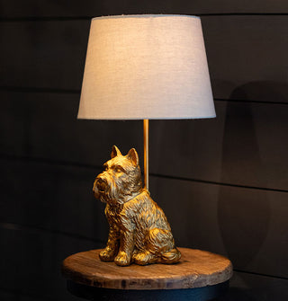 Table lamp with a beige shade and a sculptural base of a gold Scottish terrier rests on a wooden tabletop