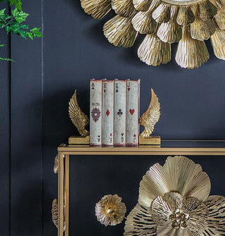 Set of metallic gold wing bookends with rectangular bases holds four books with playing card spines upright against a dark blue wall decorated with other gold items