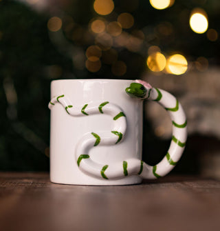 White coffee mug features a three-dimensional green striped snake with a pink flower on its head slithering across the surface and forming a handle at one side
