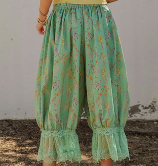Model facing away from the camera wears a pair of green floral print pantaloons with lace trim and an elastic waist