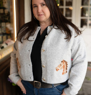Model with thumbs hooked in jeans pockets wears a gray long-sleeved button-down cardigan with brown horse details on either side near the bottom hem
