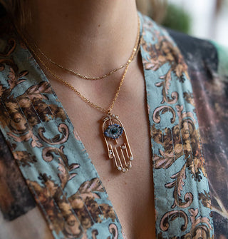 Model in a sumptuously patterned robe wears a gold wire-style hamsa hand pendant with blue and white crystals at different placements across it on gold chain