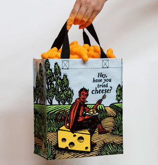 Model's hand covered in orange residue holds the black straps of a tote bag that's filled with cheese curls and features an all-over scene of a red devil sitting on a block of Swiss cheese in a pastoral setting eating cheese from a plate near the caption, "Hey, have you tried cheese?"