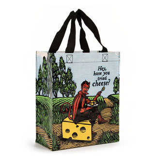 Tote bag with black straps features all-over scene of a red devil sitting on a block of Swiss cheese in a pastoral setting eating cheese from a plate near the caption, "Hey, have you tried cheese?"