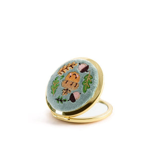 Opened round compact mirror with gold rim has a light blue velvet cover with embroidered design of a smiling acorn encircled by other smaller acorns and leaves