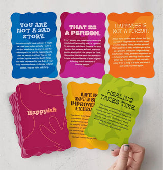 Model's hand holds a "Healing takes time" card above four others and the remaining deck of Happyish oracle cards, all brightly colored and with wavy edges