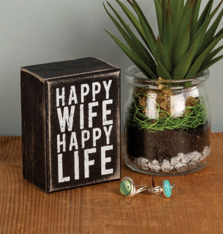 Happy Wife Happy Life box sign is staged with a potted succulent and two silver rings with green and blue gemstones on a wooden tabletop