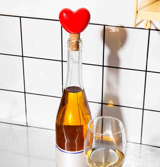 Red ceramic heart and cork bottle stopper is placed in the openeing of a wine bottle alongside a partially filled glass against a white tiled backdrop