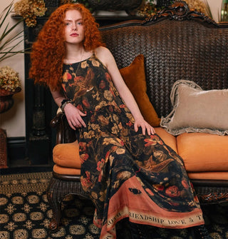 Model reclining on antique sofa wears a dark sleeveless full-length dress with lush floral print and a banner at the bottom that reads, "Friendship, love & truth"