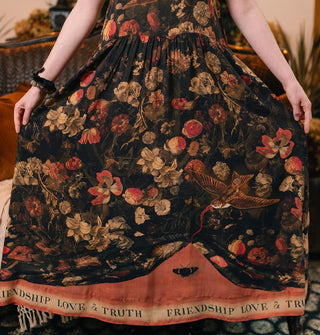 Model pulls open the skirt of a dark full-length dress with lush floral print accented by a swallow tugging at the bottom of what appears to be a tapestry, revealing a banner along the bottom hem that reads, "Friendship, love & truth"