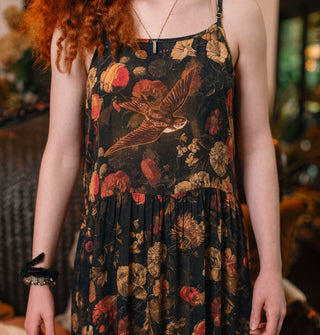 Model wears a dark sleeveless dress with lush floral print and a swallow in flight on the bodice