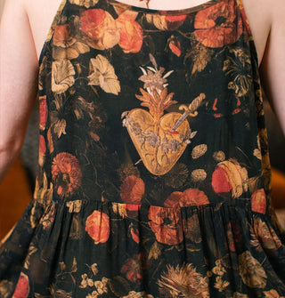 Model facing away from the camera wears a dark sleeveless dress with lush floral print and a central sacred heart pierced with a sword on its rear bodice