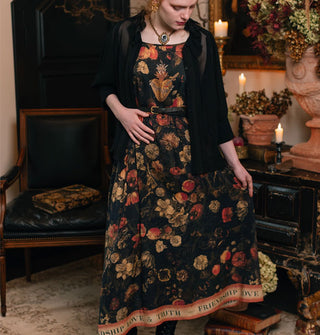 Model wears a dark full-length dress with lush floral print and the words, "Friendship, love & truth" in a banner along the bottom hem, styled with a black button-down blouse, slender belt, and antique cameo necklace
