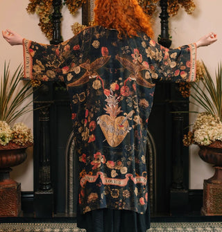 Model with arms outstretched wears a long open-front robe with dark floral print accented with banner graphics, swallows, a central heart pierced by a dagger, and border that says, "Friendship, Love & Truth"