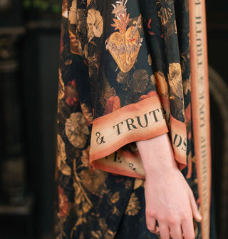Model's arm emerges from the wide sleeve of a silky robe in a dark floral and pierced heart design with contrasting border that says, "Friendship, Love & Truth"