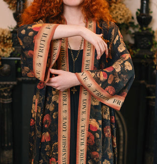 Model with arms upraised wears a long open-front robe with dark floral print accented with banner graphics and border that says, "Friendship, Love & Truth"
