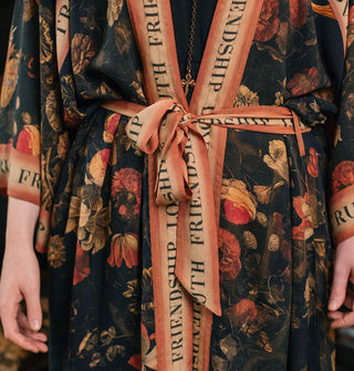 Model wears a dark floral print robe with border and belt that say "Friendship, Love & Truth"