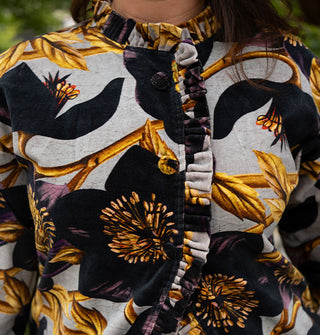 Model wears an ornately floral patterned jacket with densely pleated and ruffled edges and cloth-covered buttons