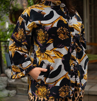 Model poses with hand in the pocket of an ornately floral patterned jacket with densely pleated and ruffled edges and cloth-covered buttons