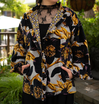 Model poses with hands in the pockets of an ornately floral patterned jacket with densely pleated and ruffled edges, cloth-covered buttons, and a contrasting printed lining