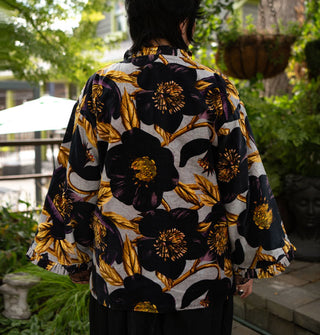 Model with back to camera wears an ornately floral black and gold on gray patterned jacket with densely pleated and ruffled neck and sleeve cuffs
