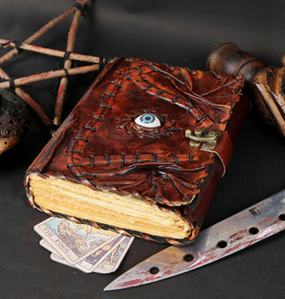 Brown leather book with rough-edged pages features textural cover details and stitching, a central blue eye, and strap closure with brass lock is staged with a bloodied knife, tarot cards, and a handmade wooden star