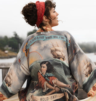 Model wears a loose-fitting robe with classical seascape print and text banner artwork that says, "Smooth seas & fair winds will find you — hold fast"
