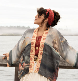 Model leaning against a wooden railing in front of a misty lakeside backdrop wears a loose-fitting cropped robe with classical seascape artwork and a contrasting orange moon and sun print trim
