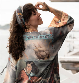 Model with hand raised to forehead wears a loose-fitting silken robe featuring classical seascape artwork with text banner accents and a contrasting orange sun and moon print sleeve trim