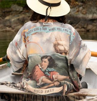 Model rowing a boat and facing away from the camera wears a loose-fitting silken robe that features classical seascape artwork with text banner accents that read, "Smooth seas & fair winds will find you — hold fast"