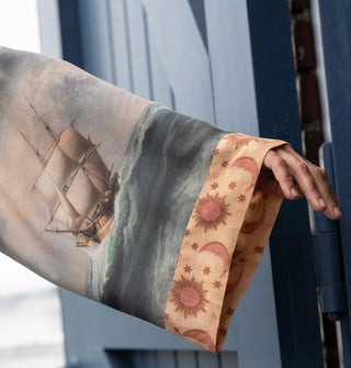 Model's outstretched arm showcases a loose-fitting silken robe sleeve with classical brig artwork and a contrasting orange sun and moon trim