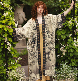 Model standing in a garden setting wears a lightweight robe with black and white floral design contrasted by bee print borders