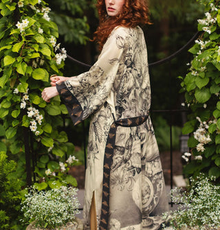 Model standing in a garden setting wears a lightweight robe with black and white floral design contrasted by bee print cuffs and belt