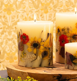 Lit pillar candles with flowers and leaves embedded in the wax are displayed on a wooden platform against a yellow and white damask backdrop