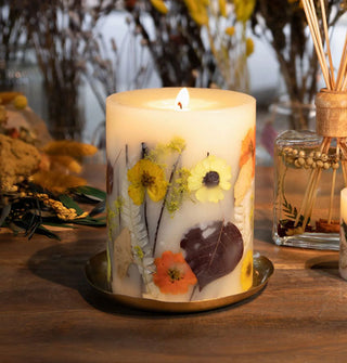 Lit pillar candle with colorful flowers and leaves embedded in the wax rests on a wooden surface decorated with a reed diffuser and other dried botanicals