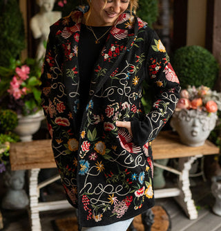 Smiling model standing against a backdrop of potted plants and a bench on a porch deck wears a long black cardigan with colorful embroidered designs and the words, "Hope" and "Love" in white script all over it
