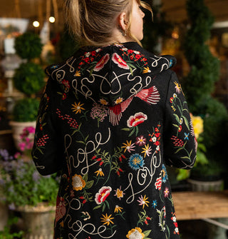 Model standing against a backdrop of potted plants with back to camera wears a black hooded cardigan with colorful embroidered designs and the words, "Hope" and "Love" in white script all over it