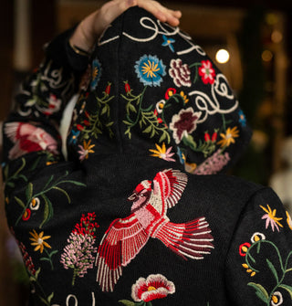 Model with back to camera wears the hood of a black cardigan with colorful embroidered floral and bird designs and white script all over it