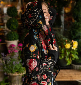 Smiling model standing against a backdrop of potted plants wears a black hooded cardigan with colorful embroidered designs and white script all over it